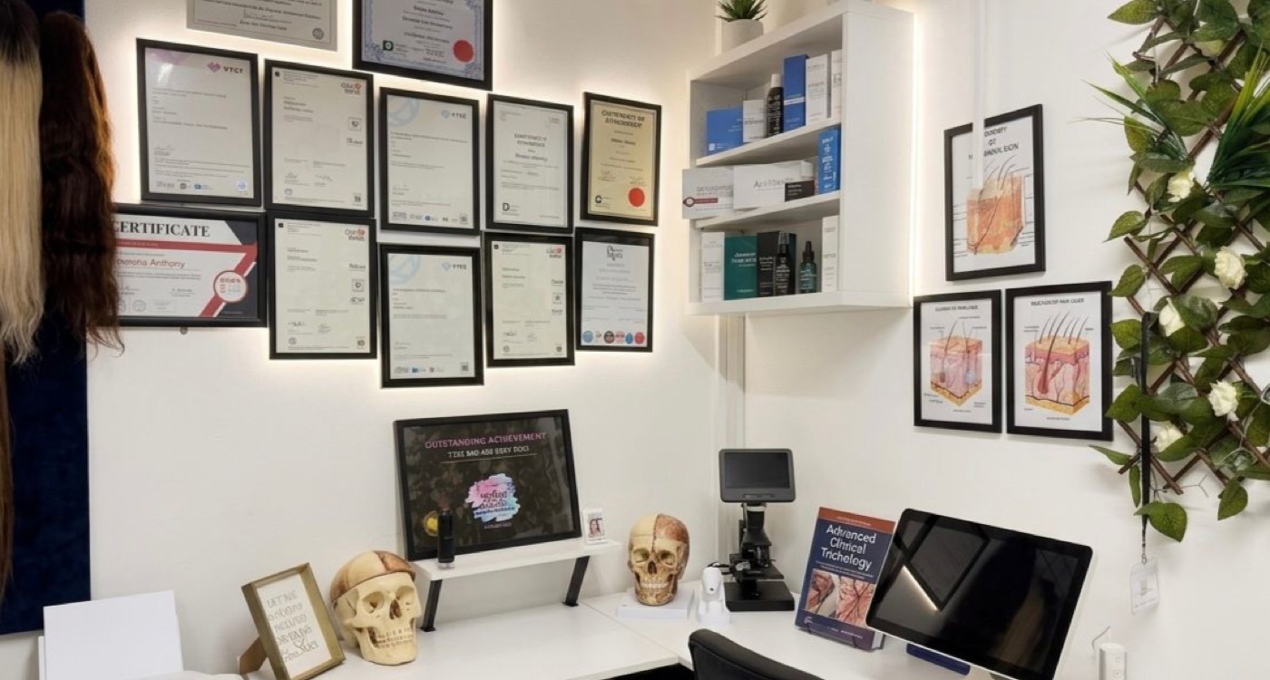 A wall of framed trichology certificates in the clinic's consultation room, beside a desk holding a digital trichoscope, anatomical hair-follicle diagrams and a clinical trichology reference text.